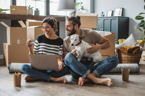 Casal jovem a mudar-se para a nova casa. Estão felizes e a usar o portátil para comprar o que precisam.